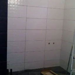 Wnętrze łazienki w trakcie remontu: białe płytki ścienne z subtelnym wzorem, czarne płytki podłogowe, zabezpieczona folią podłoga, widoczne otwory na instalacje, drabina oparta o ścianę.