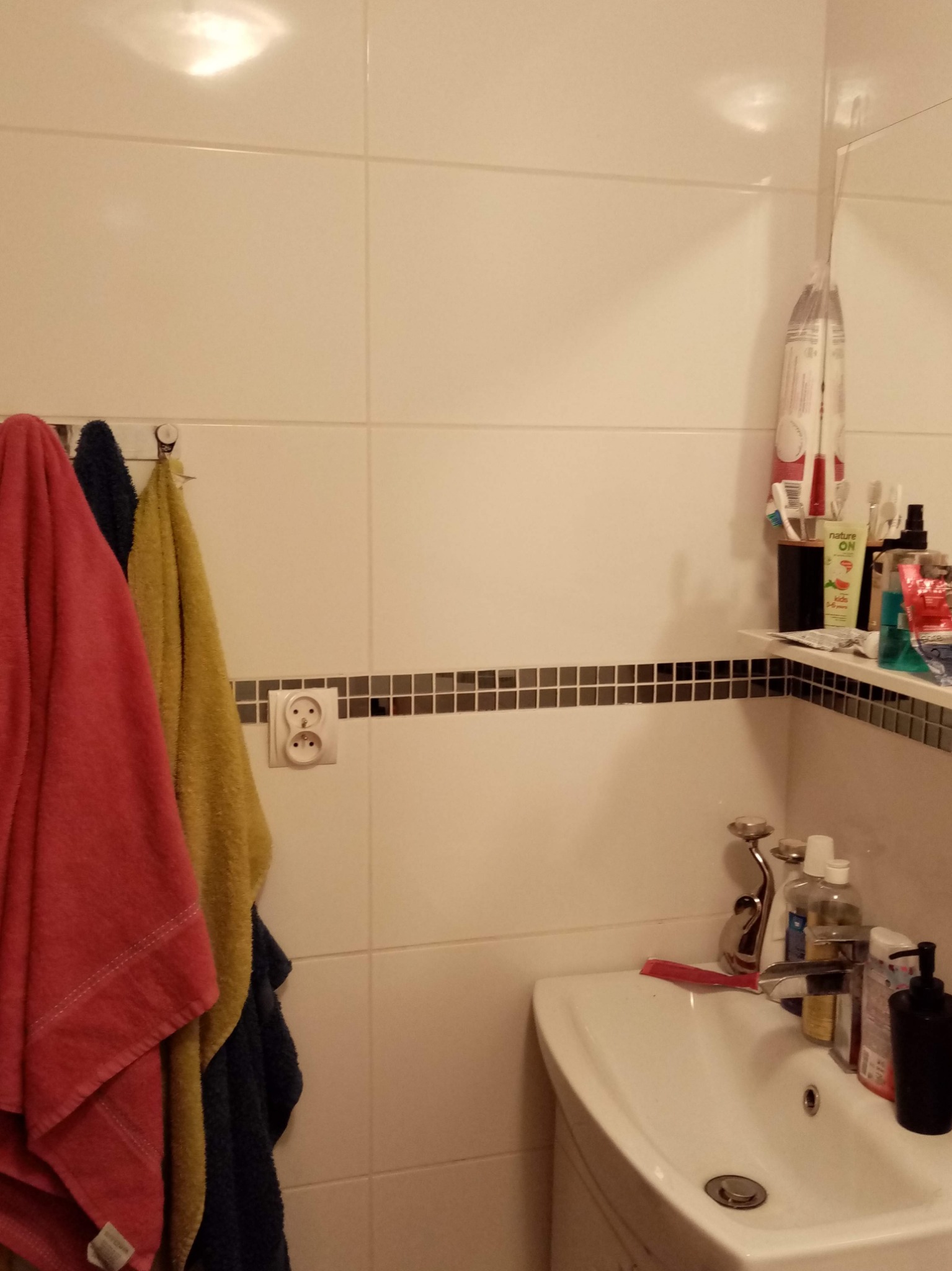 Łazienka z umywalką, płytkami w odcieniach bieli i beżu, dekoracyjnym paskiem z mozaiki, wiszącymi ręcznikami w kolorach: różowym, żółtym i granatowym oraz gniazdkiem elektrycznym na ścianie.