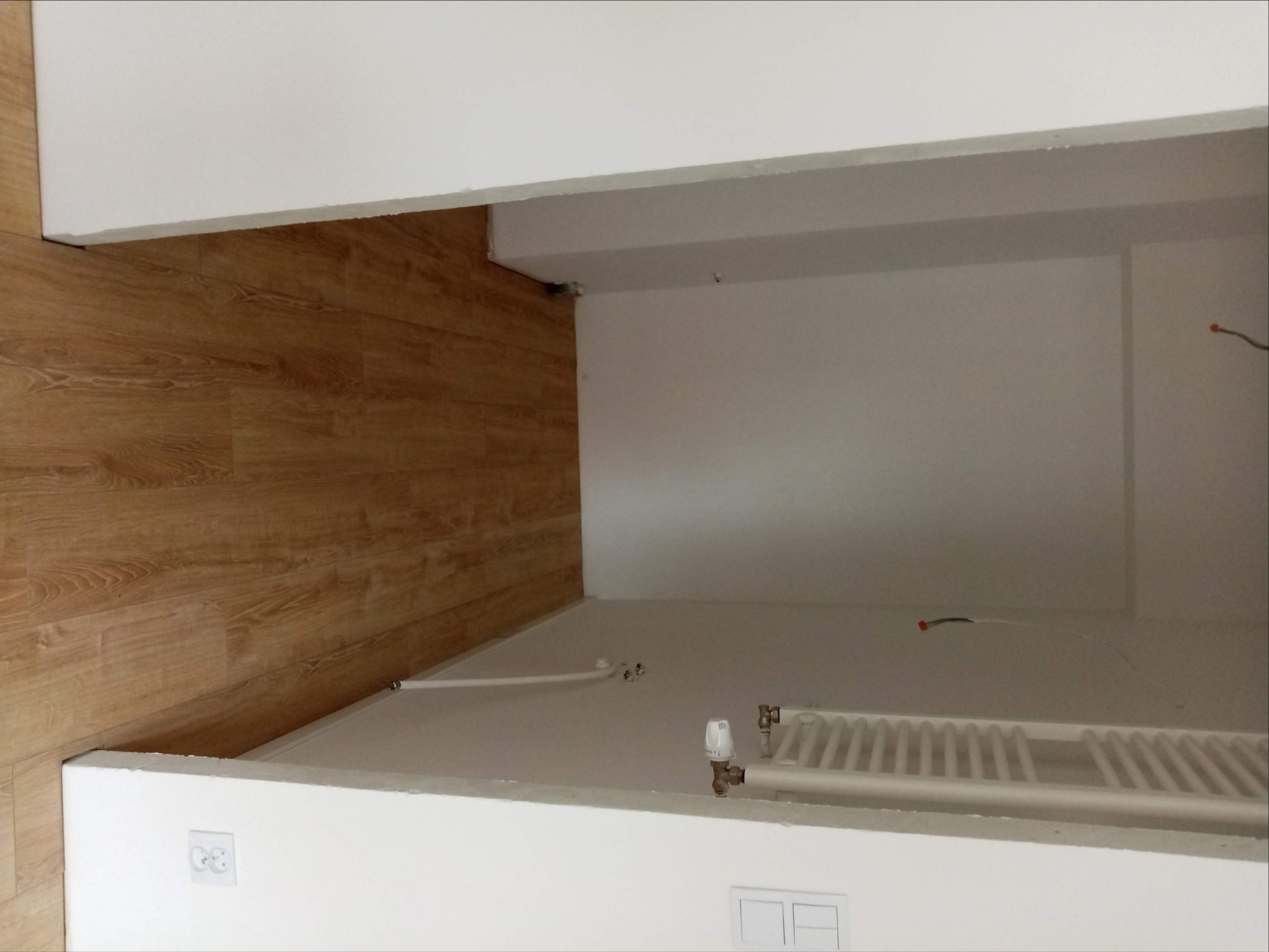 Surowe wykończenie wnętrza z drewnianą podłogą, białą ścianą, grzejnikiem i wystającymi kablami elektrycznymi przed malowaniem.