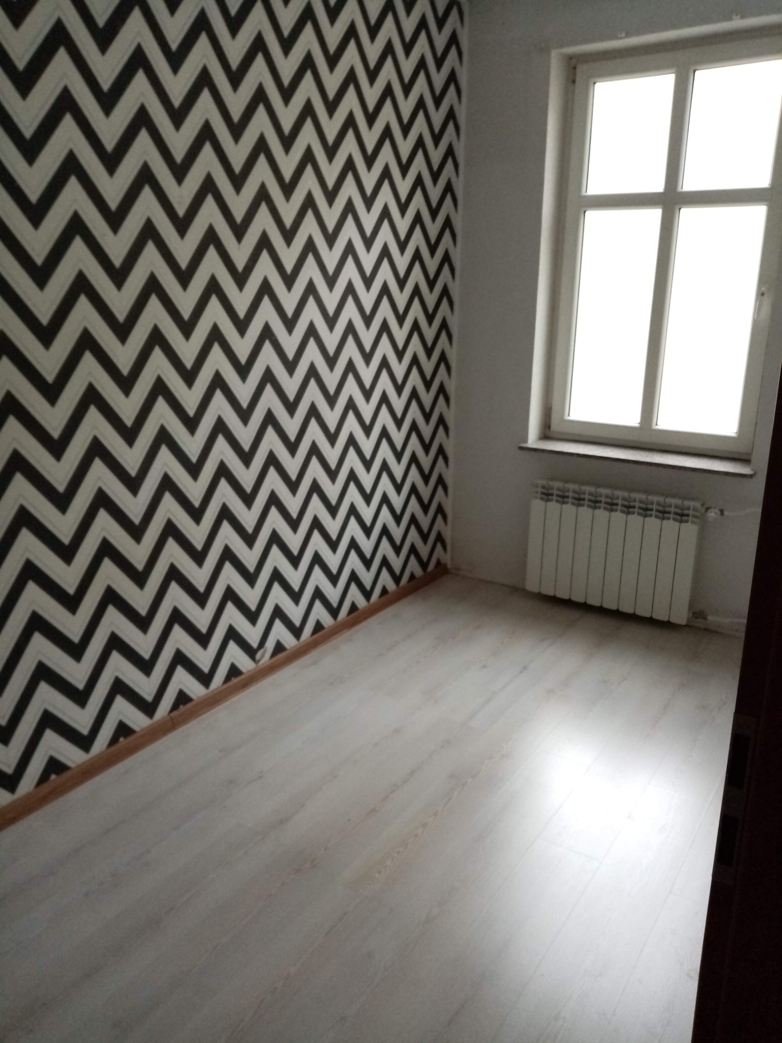 Pokój po remoncie z białą podłogą, ścianą w zygzaki i białym grzejnikiem pod oknem.