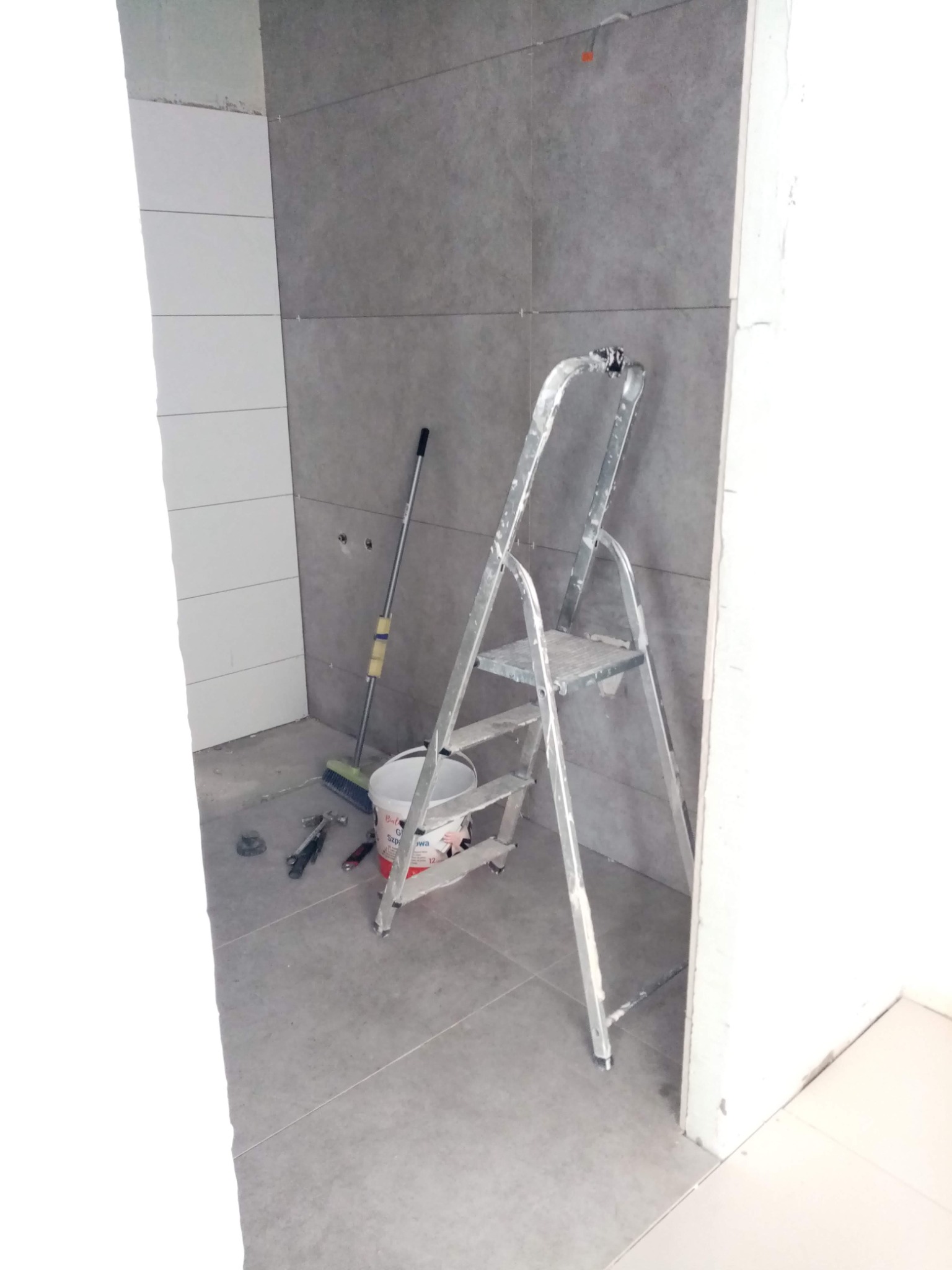 Świeżo ułożone szare płytki na ścianie i podłodze w łazience w Ełku, z aluminiową drabiną, wiadrem i narzędziami glazurniczymi w tle.