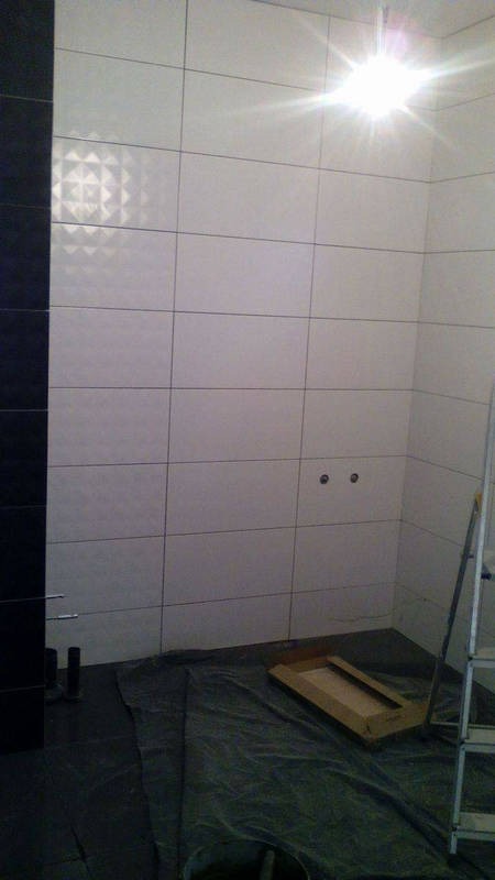 Wnętrze łazienki w trakcie remontu: białe płytki ścienne z subtelnym wzorem, czarne płytki podłogowe, zabezpieczona folią podłoga, widoczne otwory na instalacje, drabina oparta o ścianę.