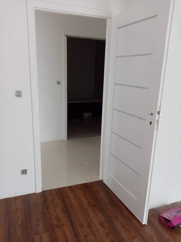 Otwarta biała, lakierowana, wewnętrzna stolarka drzwiowa z poziomymi frezowaniami, widoczna na tle wykafelkowanego pomieszczenia z jasnymi płytkami podłogowymi, oraz brązową podłogą drewnopodobną...