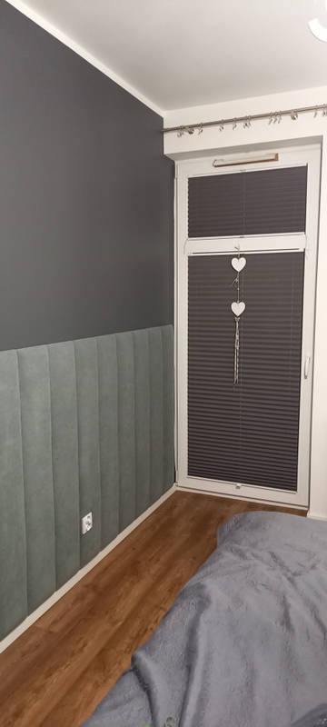 Szara ściana z panelami tapicerowanymi w pionowe pasy, drewniana podłoga, okno z plisowanymi roletami i dekoracją w kształcie serc.