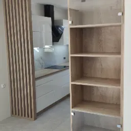 Wysoka, wąska szafka z półkami w odcieniach drewna, częściowo przeszklona, na tle nowoczesnej, białej kuchni z drewnianym blatem i czarnym okapem, widoczna także drewniana ścianka działowa...