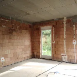 Surowy stan wnętrza z widoczną instalacją elektryczną: kable prowadzone po ścianach z czerwonej cegły i pod sufitem, przygotowane punkty pod gniazdka i włączniki, okna z widokiem na zieleń.