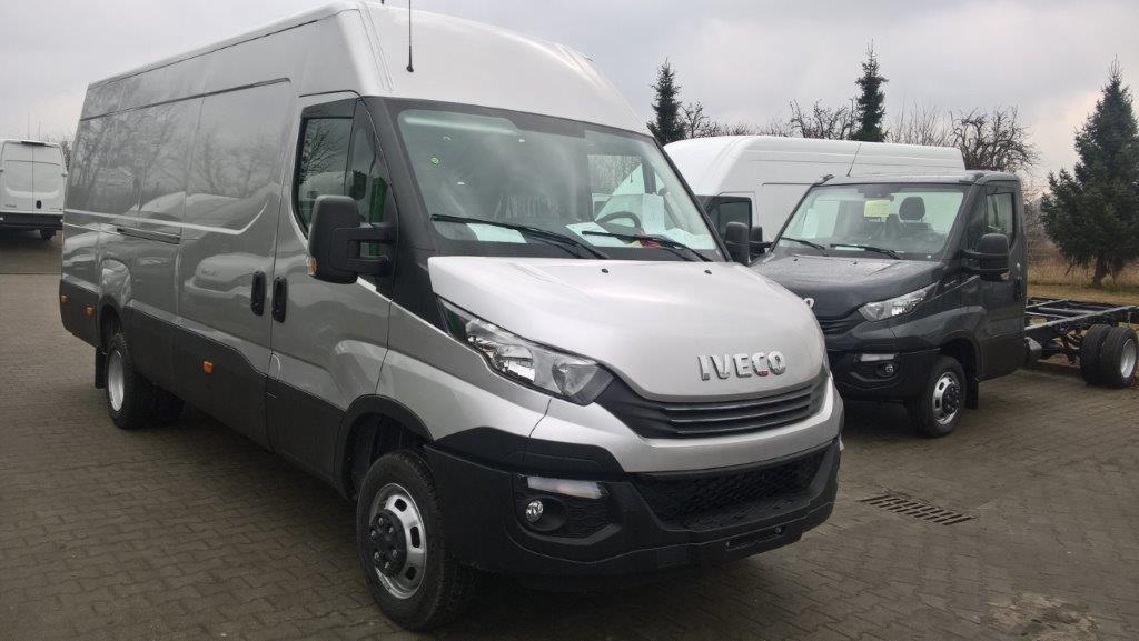 Srebrny bus Iveco z wysokim dachem na placu, z widocznymi innymi pojazdami w tle.