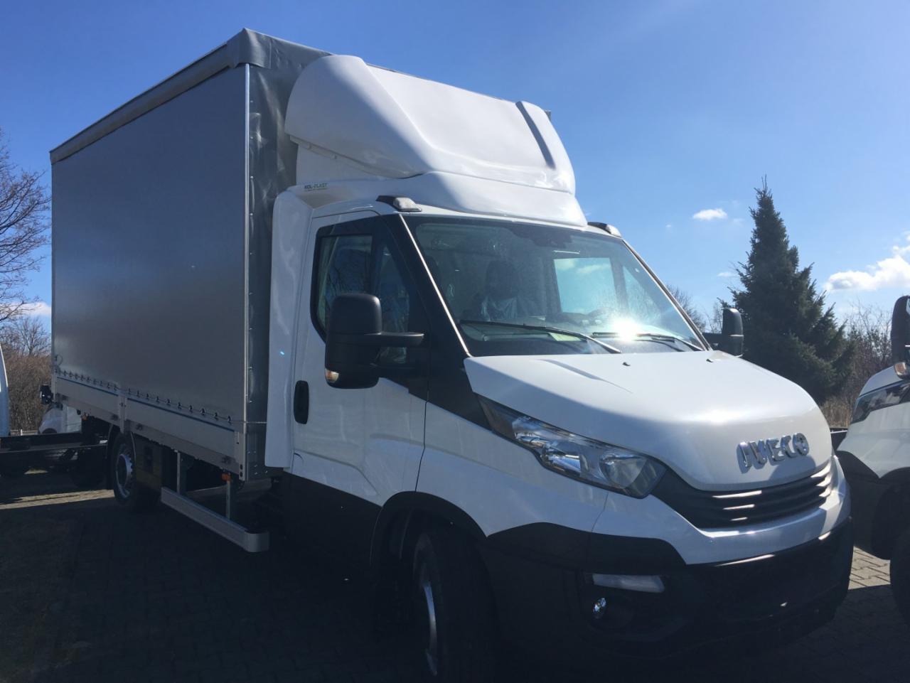 Biały samochód dostawczy marki Iveco z szarą plandeką na parkingu, widoczne drzewa w tle, niebieskie niebo.