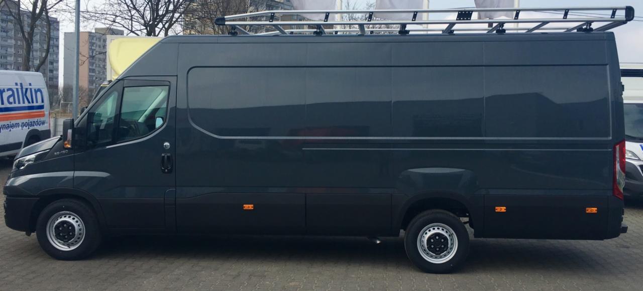 Ciemnoszary bus z relingami dachowymi zaparkowany na betonowym placu, widok z boku, w tle budynki mieszkalne.