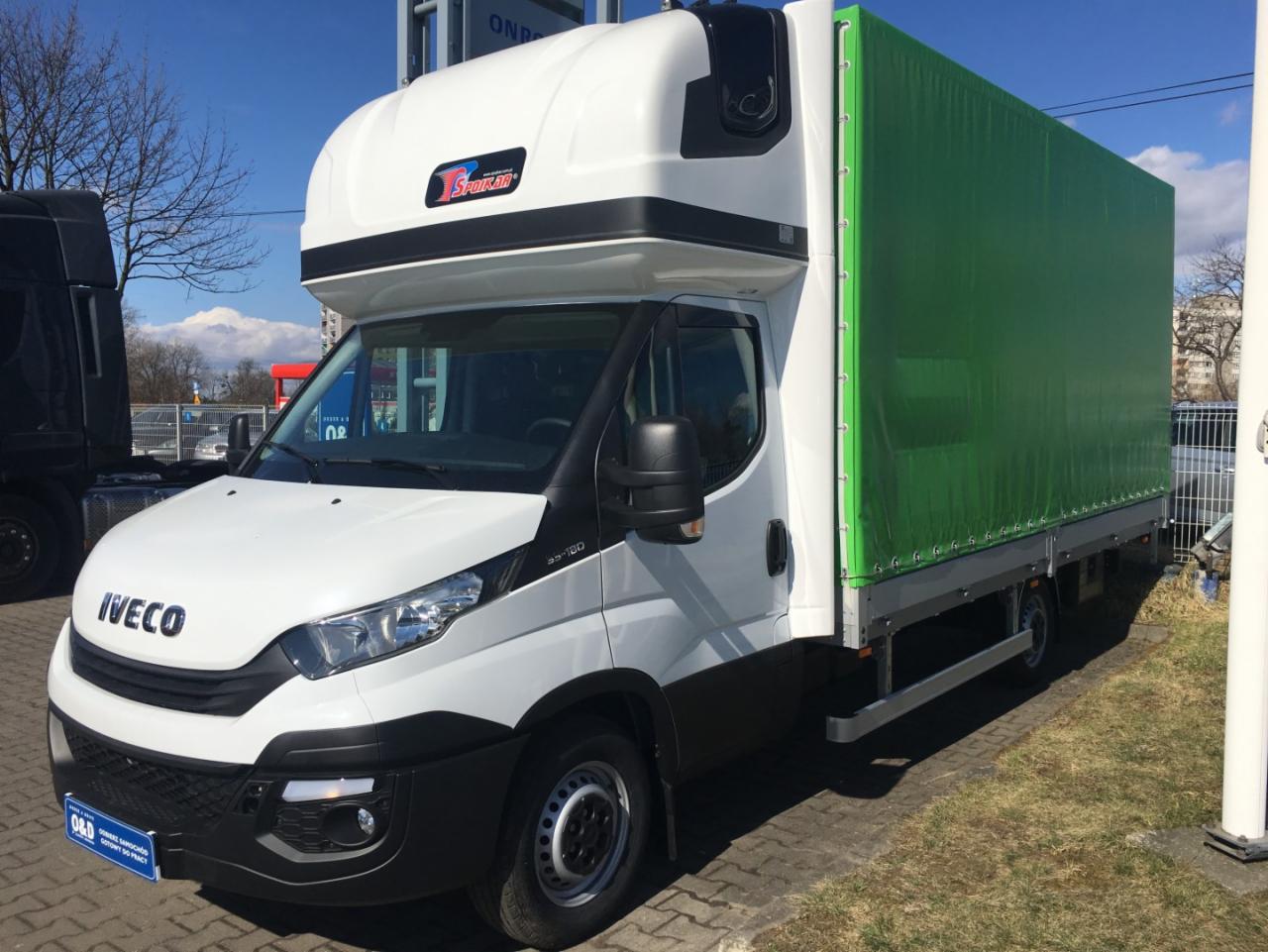 Biały samochód dostawczy Iveco z zieloną plandeką, zaparkowany na zewnątrz w słoneczny dzień.