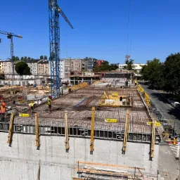 Wysoki kąt widzenia na plac budowy z pracującymi robotnikami na zbrojeniu, betonowy fundament w trakcie budowy, w tle budynki mieszkalne i żurawie budowlane.