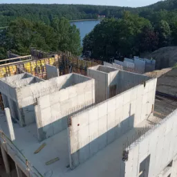 Wznoszenie ścian żelbetowych budynku mieszkalnego z widocznym zbrojeniem, szalunkami i malowniczym jeziorem w tle, uchwycone z góry w słoneczny dzień.