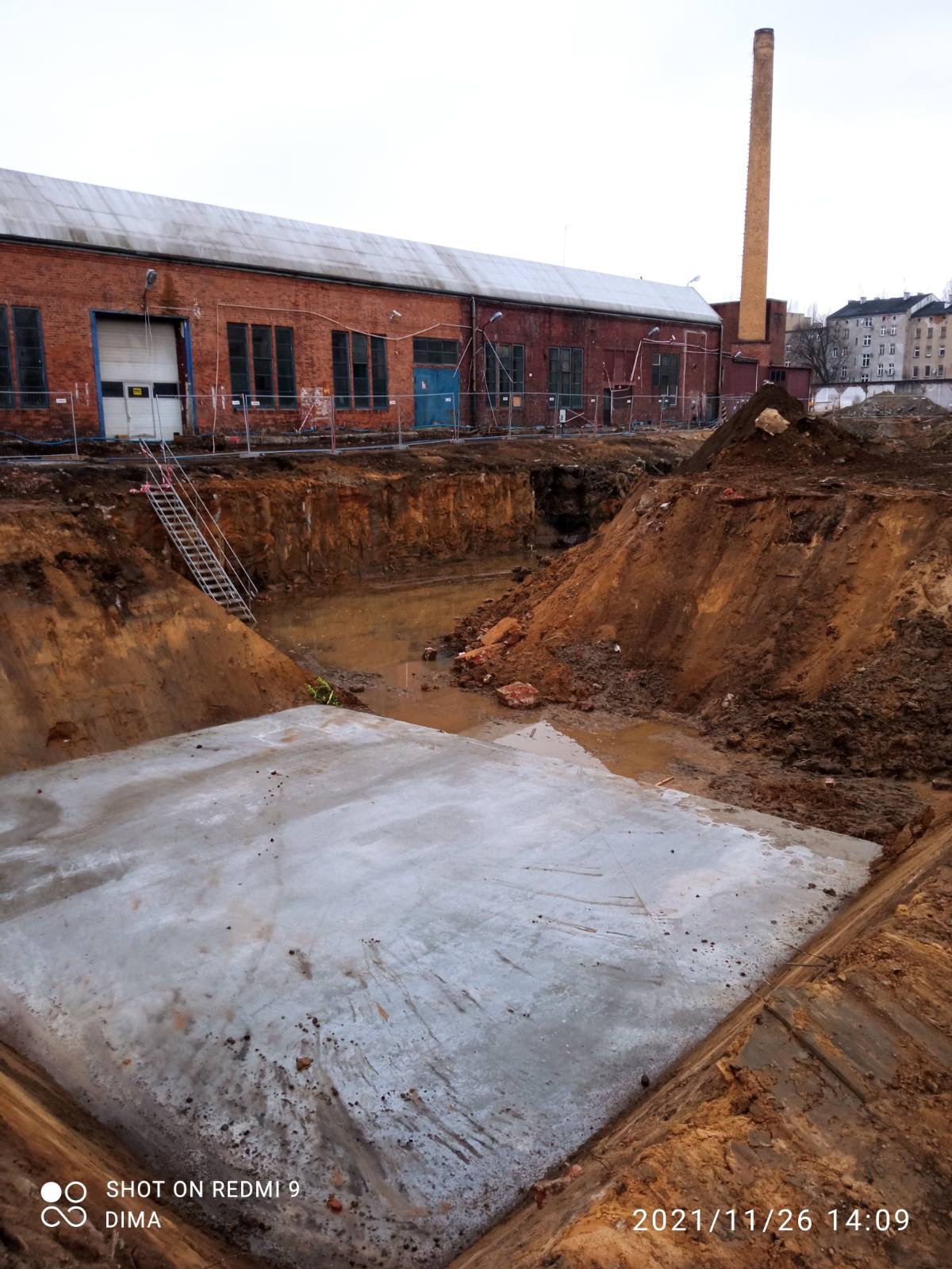 Wykop z wylaną betonową płytą fundamentową na terenie budowy, widoczny budynek z czerwonej cegły w tle.