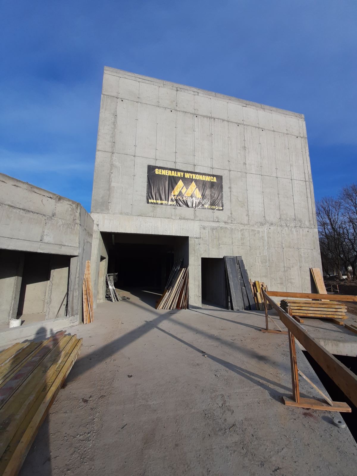 Surowy budynek żelbetowy w trakcie budowy, z widocznym banerem 'Generalny Wykonawca', drewniane stemple i deski na placu budowy, błękitne niebo w tle.