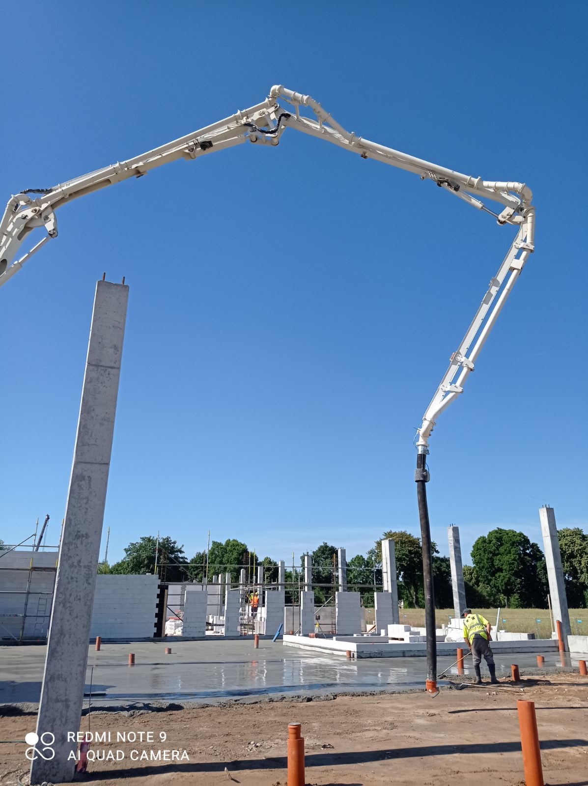 Betonowanie fundamentów domu jednorodzinnego z użyciem pompy do betonu, widoczne słupy i ściany z betonu komórkowego, pracownik w żółtej kamizelce wyrównuje powierzchnię betonu.