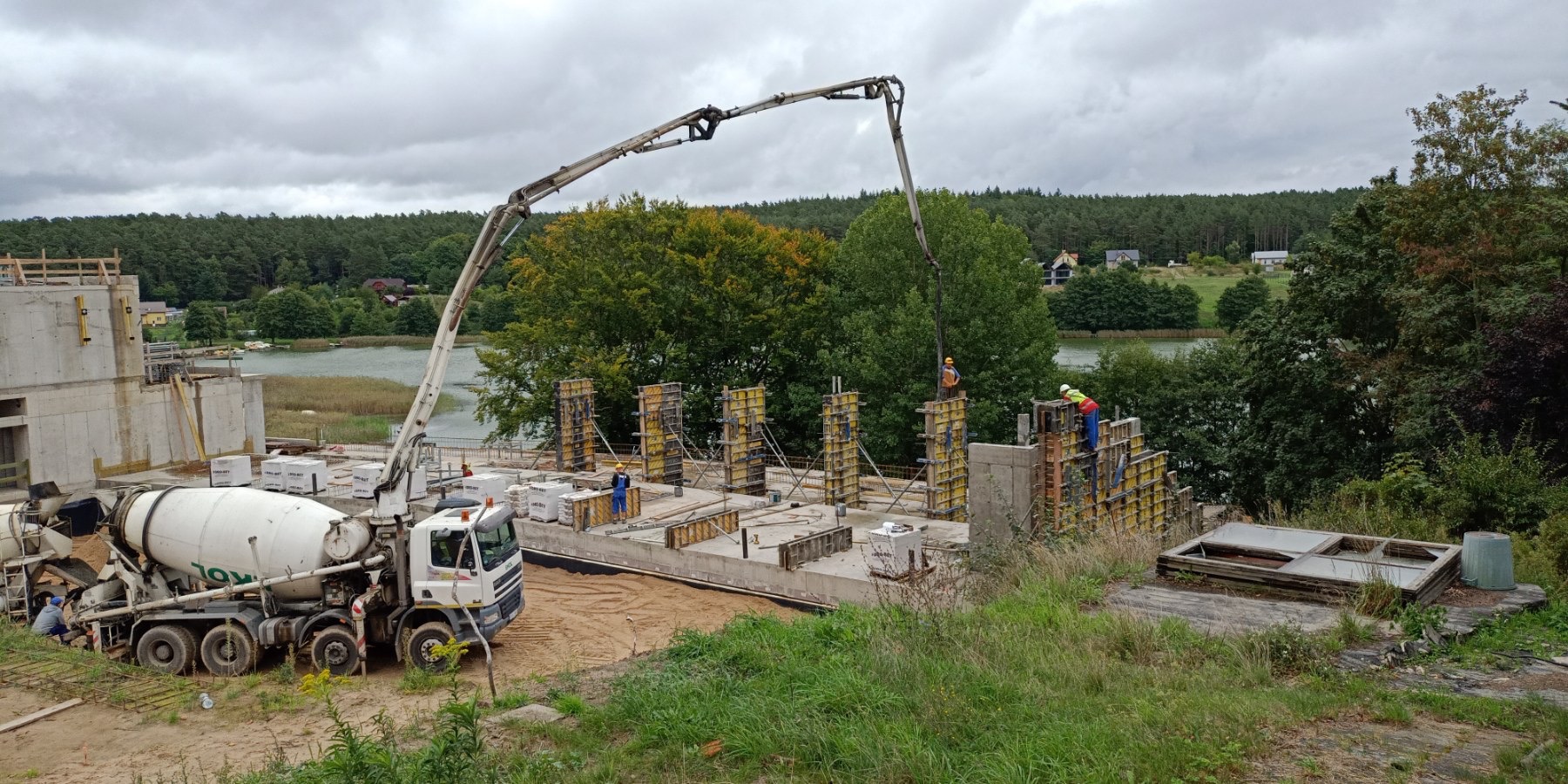 Wylewanie betonu na fundamenty budynku przy użyciu pompy do betonu, widoczny mikser do betonu, szalunki, pracownicy w kaskach i odblaskowych kamizelkach, jezioro i drzewa w tle.
