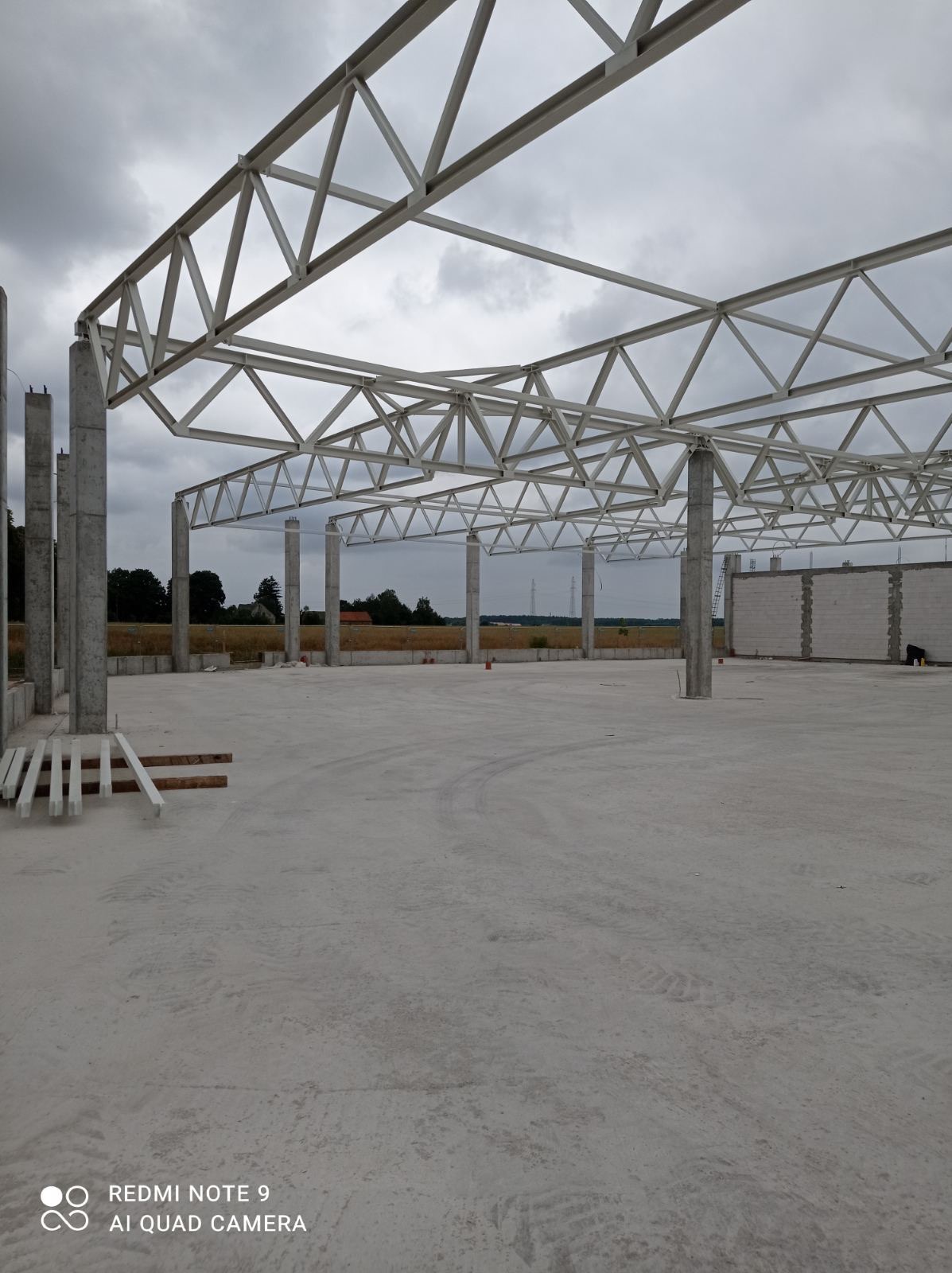 Wnętrze hali w trakcie budowy: betonowa posadzka, żelbetowe słupy podtrzymujące stalową konstrukcję dachu, widoczne bloczki betonowe na ścianie w tle.