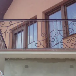 Kuta, metalowa balustrada balkonowa z dekoracyjnymi zawijasami, zamontowana przed oknami w budynku o pomarańczowej elewacji.