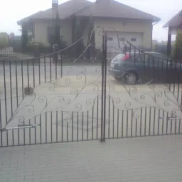 Kuta brama wjazdowa z automatycznym napędem, zdobiona ornamentami roślinnymi, na tle posesji z domem i zaparkowanym samochodem.