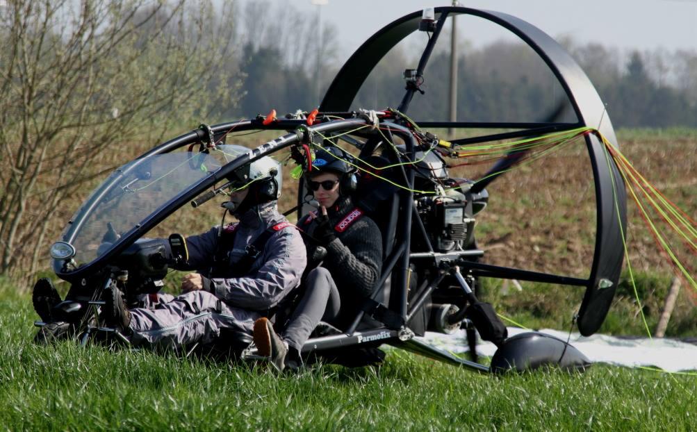 Dwóch pilotów siedzi w ultralekkim samolocie na trawiastym polu, jeden z nich pokazuje znak 'V' palcami.