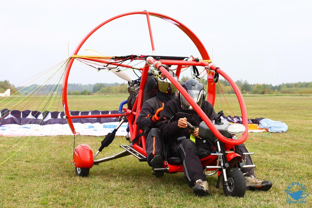 Dwuosobowy motolotniarz w czerwonej ramie z silnikiem, siedzący na trawiastym lotnisku z rozłożonym spadochronem w tle, przygotowujący się do startu.