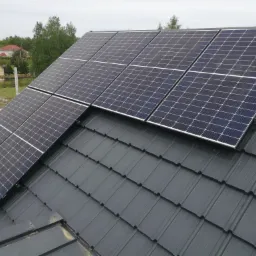 Panele słoneczne zamontowane na ciemnoszarym dachu z dachówki ceramicznej, widok z góry, w tle zieleń drzew i zabudowania mieszkalne.