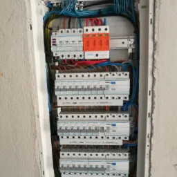 Rozdzielnica elektryczna Hager w białej obudowie, widoczne zabezpieczenia Simetec, przewody w różnych kolorach oraz fragment ściany w surowym stanie.