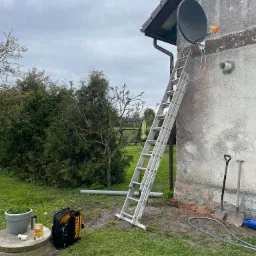 Szara elewacja budynku z zamontowaną anteną satelitarną, drabina oparta o ścianę, narzędzia i skrzynka z osprzętem na trawniku, pochmurne niebo.