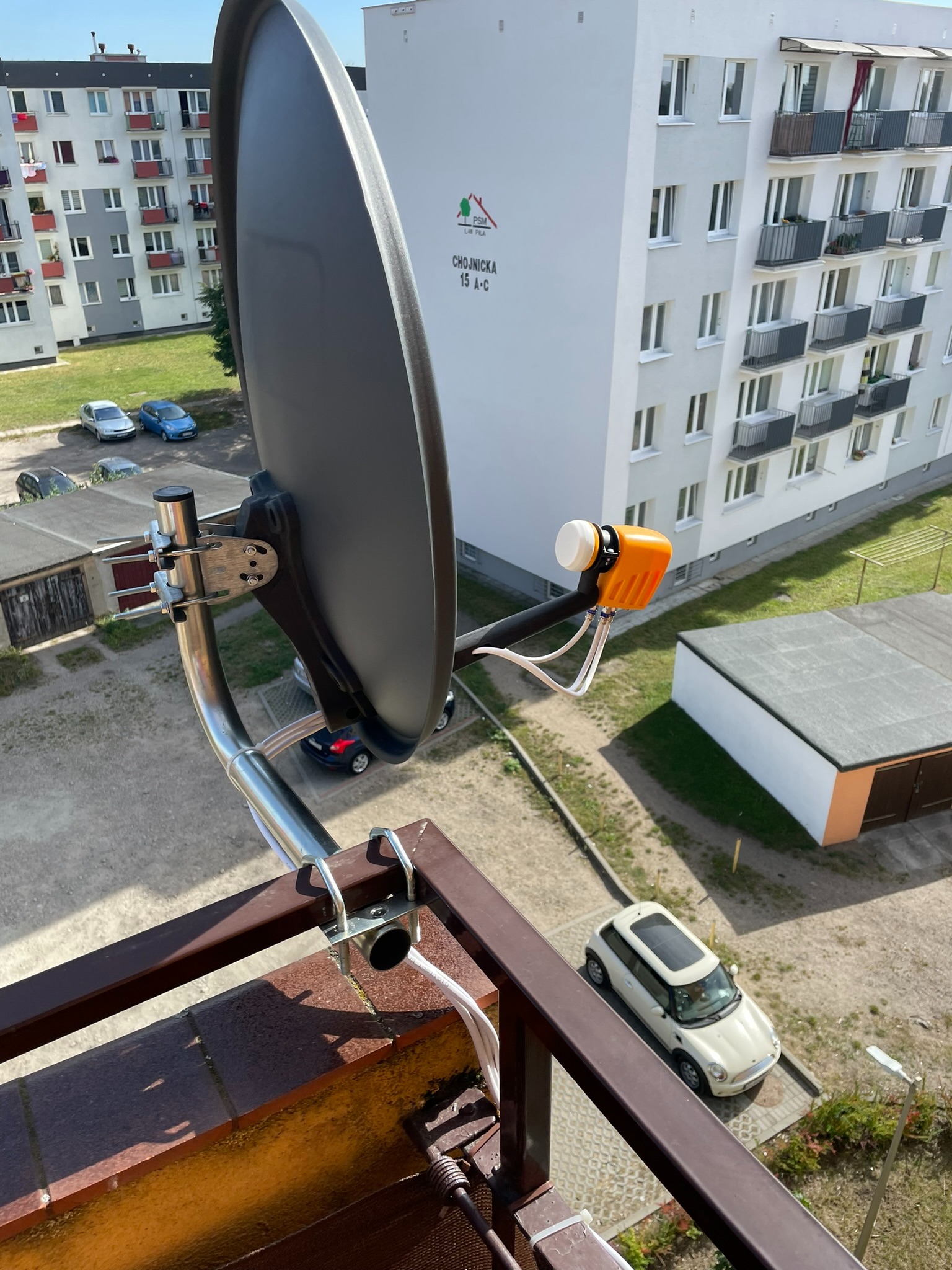 Szara antena satelitarna zamontowana na balkonie bloku mieszkalnego, widok z góry na okolicę.