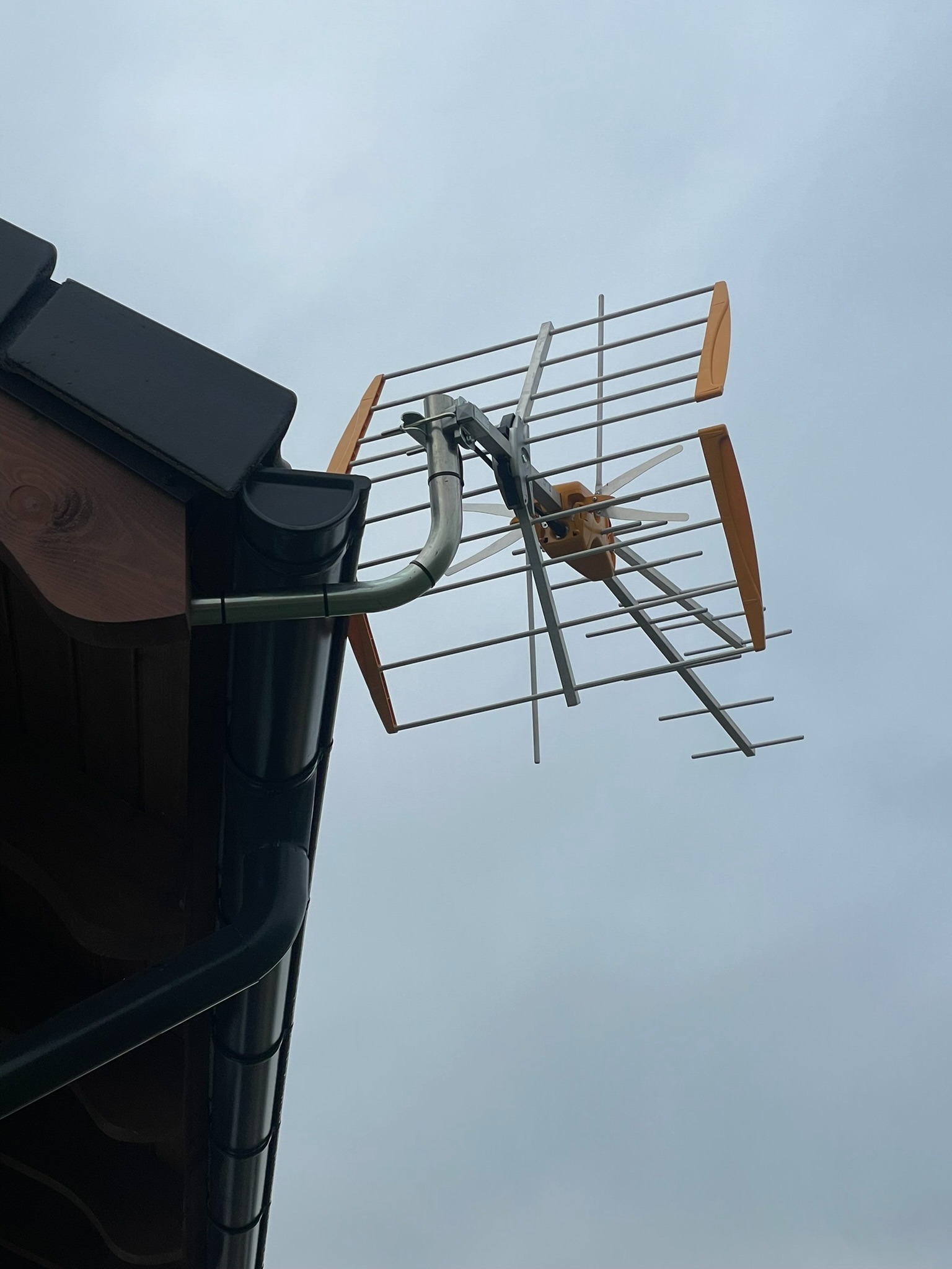 Antena telewizyjna zamontowana na dachu domu, przymocowana do rynny za pomocą metalowego uchwytu, na tle pochmurnego nieba.