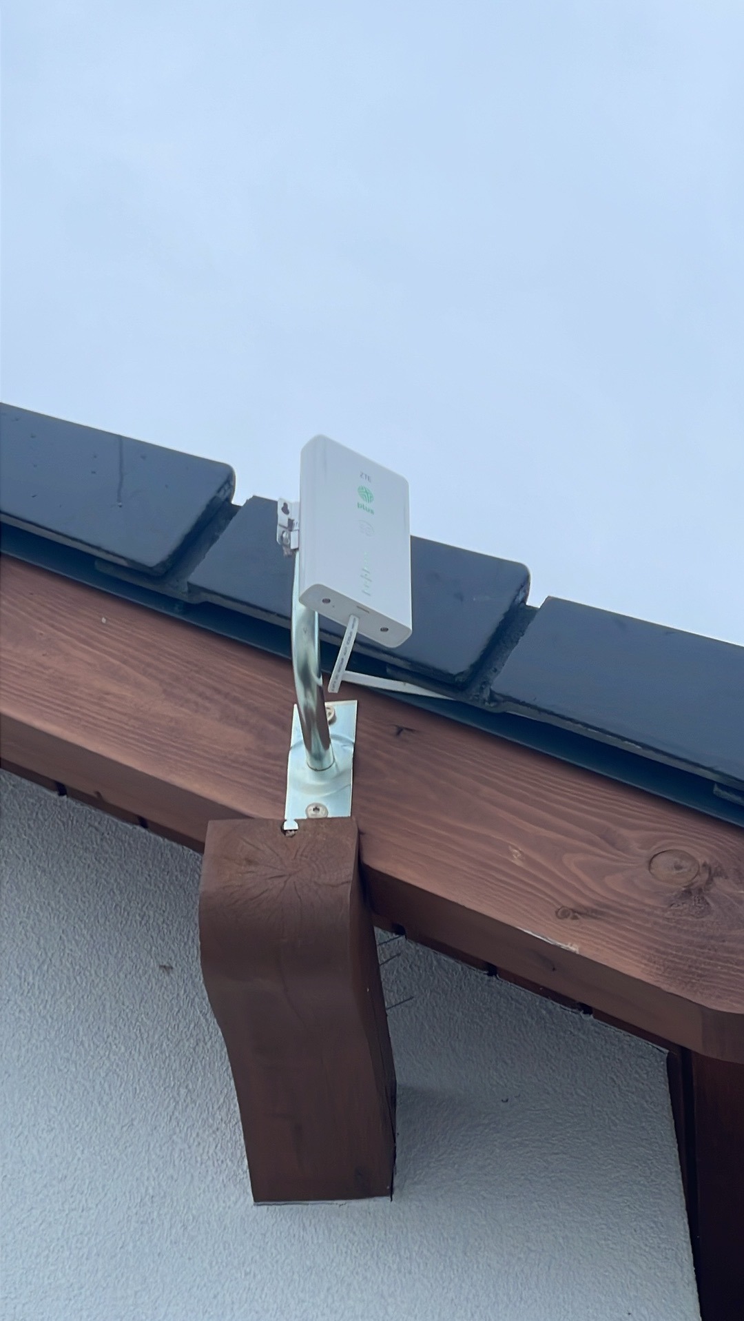 Zewnętrzna, biała antena ZTE zamocowana na metalowym uchwycie do drewnianej belki pod dachem z ciemnej dachówki, widok od dołu.