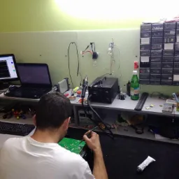 Mężczyzna w białej koszulce lutuje płytę drukowaną na biurku warsztatowym z komputerem, oscyloskopem, narzędziami i organizerem z częściami elektronicznymi w tle.