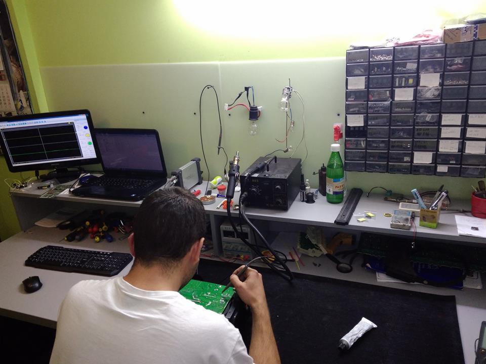 Mężczyzna w białej koszulce lutuje płytę drukowaną na biurku warsztatowym z komputerem, oscyloskopem, narzędziami i organizerem z częściami elektronicznymi w tle.