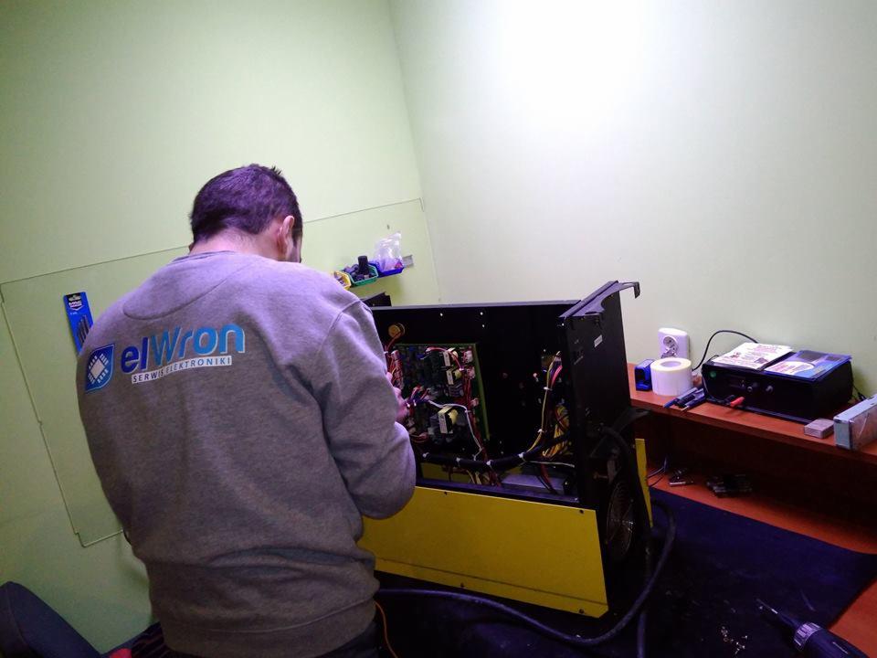 Serwisant w szarej bluzie z logo 'elWron Serwis Elektroniki' naprawia otwarty, żółto-czarny sprzęt elektroniczny z widocznymi płytkami drukowanymi w pomieszczeniu z zielonymi ścianami.