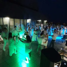 Grupa osób ubranych na biało tańczy na oświetlonym na zielono parkiecie podczas wieczornej imprezy na dachu budynku.