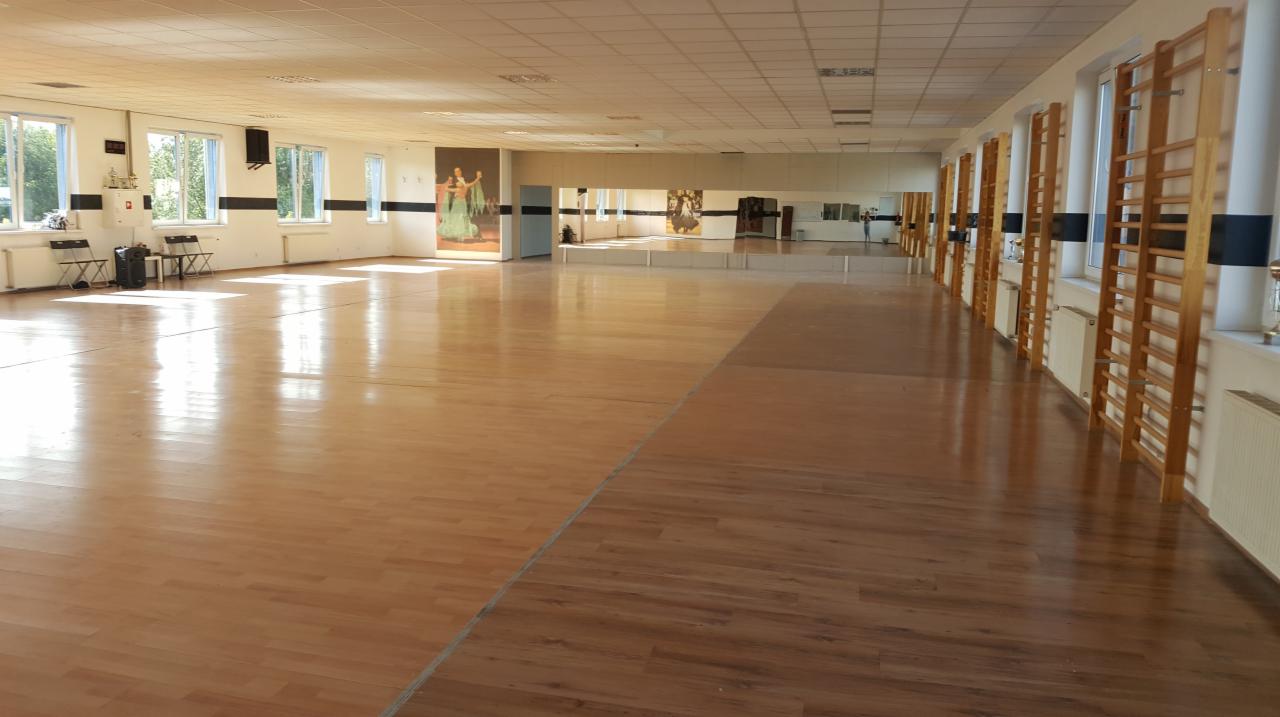 Przestronna sala taneczna z drewnianą podłogą, lustrami wzdłuż ściany i drabinkami gimnastycznymi, oświetlona naturalnym światłem z okien.