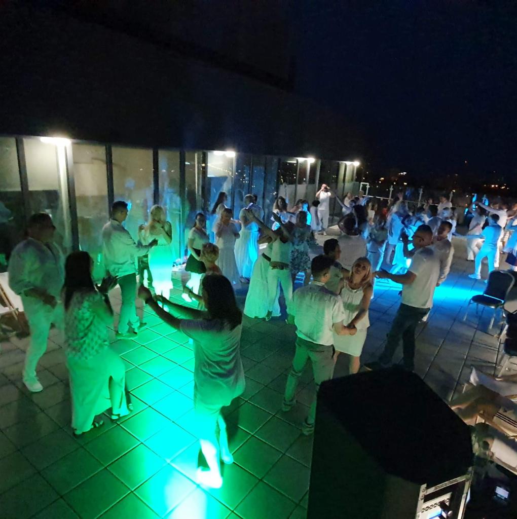 Grupa osób ubranych na biało tańczy na oświetlonym na zielono parkiecie podczas wieczornej imprezy na dachu budynku.