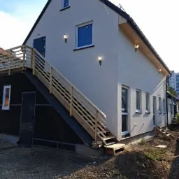 Nowoczesny dwukondygnacyjny dom z jasną elewacją, drewnianymi schodami zewnętrznymi prowadzącymi do drzwi na piętrze, oświetleniem zewnętrznym i widocznymi oknami.