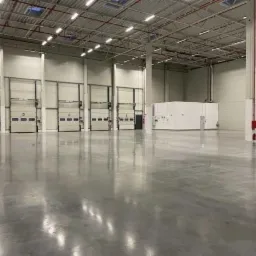 Puste wnętrze magazynu z betonową podłogą, widoczne bramy segmentowe i biały klimatyzator przemysłowy.