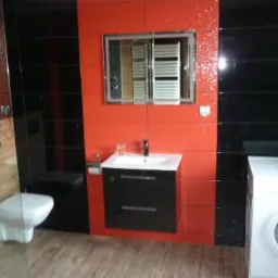 Wyremontowana łazienka z czarno-czerwonymi płytkami, umywalką z czarną szafką, lustrem z podświetleniem LED, toaletą podwieszaną i pralkami po bokach.