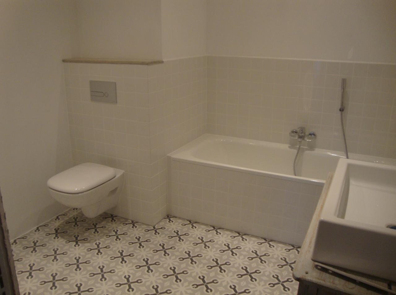 Wykończona łazienka z białą armaturą, płytkami na ścianach i wzorzystą podłogą, widok na toaletę, wannę i umywalkę.