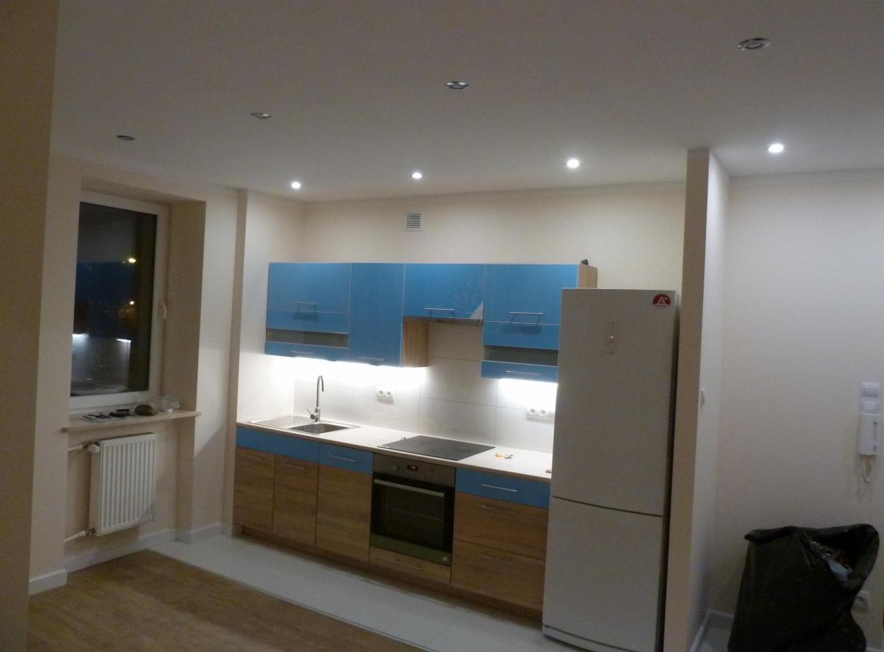 Jasna kuchnia po remoncie z błękitnymi szafkami wiszącymi i drewnopodobnymi dolnymi, oświetleniem podszafkowym LED, białym blatem i lodówką, widok na okno i grzejnik.