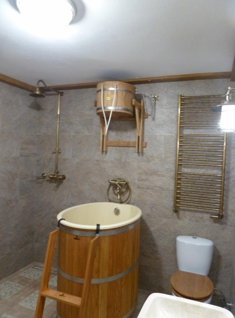 Drewniana wanna beczkowa z drabinką, prysznic retro, grzejnik drabinkowy i toaleta w łazience wykończonej płytkami imitującymi kamień.