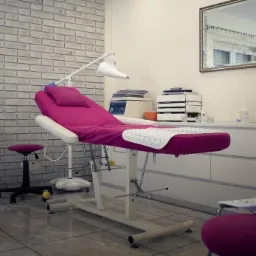 Fotel do zabiegów kosmetycznych z fuksjowym obiciem i białym podkładem z wypustkami, na tle ściany z białej cegły i białych szafek.