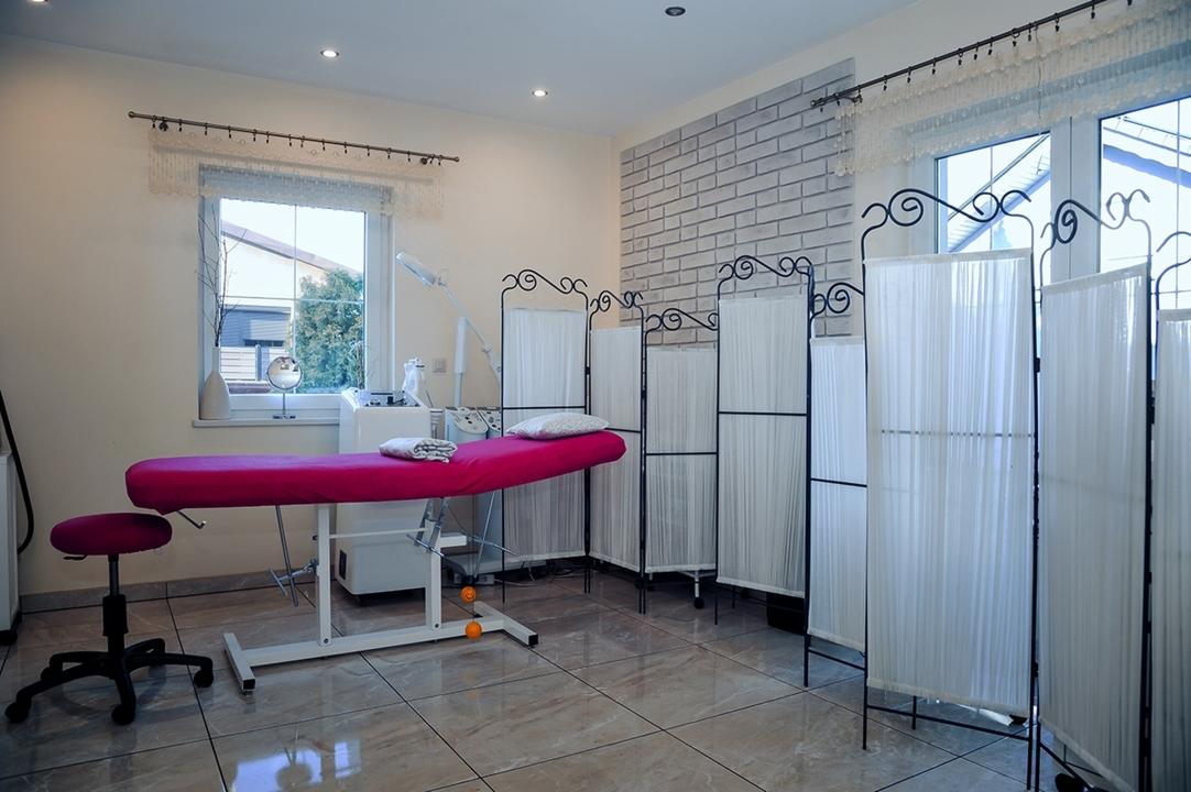 Jasne wnętrze gabinetu z fuksjowym leżankiem, parawanem z białymi zasłonami, urządzeniami kosmetycznymi i oknem z widokiem na zieleń.