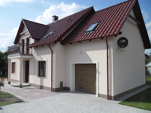 Dwukondygnacyjny dom jednorodzinny z ocieploną elewacją w odcieniach beżu, z czerwonym dachem, balkonem z ozdobną balustradą i anteną satelitarną na ścianie garażu.
