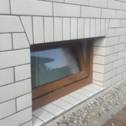 Elewacja budynku wykończona białymi płytkami imitującymi cegłę, z drewnianą ramą okienną i małym oknem uchylnym, widok z zewnątrz.