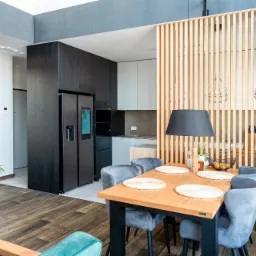 Nowoczesna jadalnia z drewnianym stołem, szarymi krzesłami i ażurową ścianką działową z jasnego drewna, widoczna kuchnia w tle.