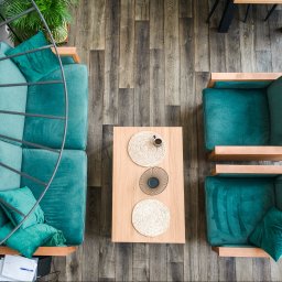 PERFECT ROOM - Widok z góry na salon z drewnianą podłogą, turkusową sofą i fotelami z drewnianymi ramami, stolik kawowy z dekoracjami i filiżanką, oraz fragment metalowej lampy.