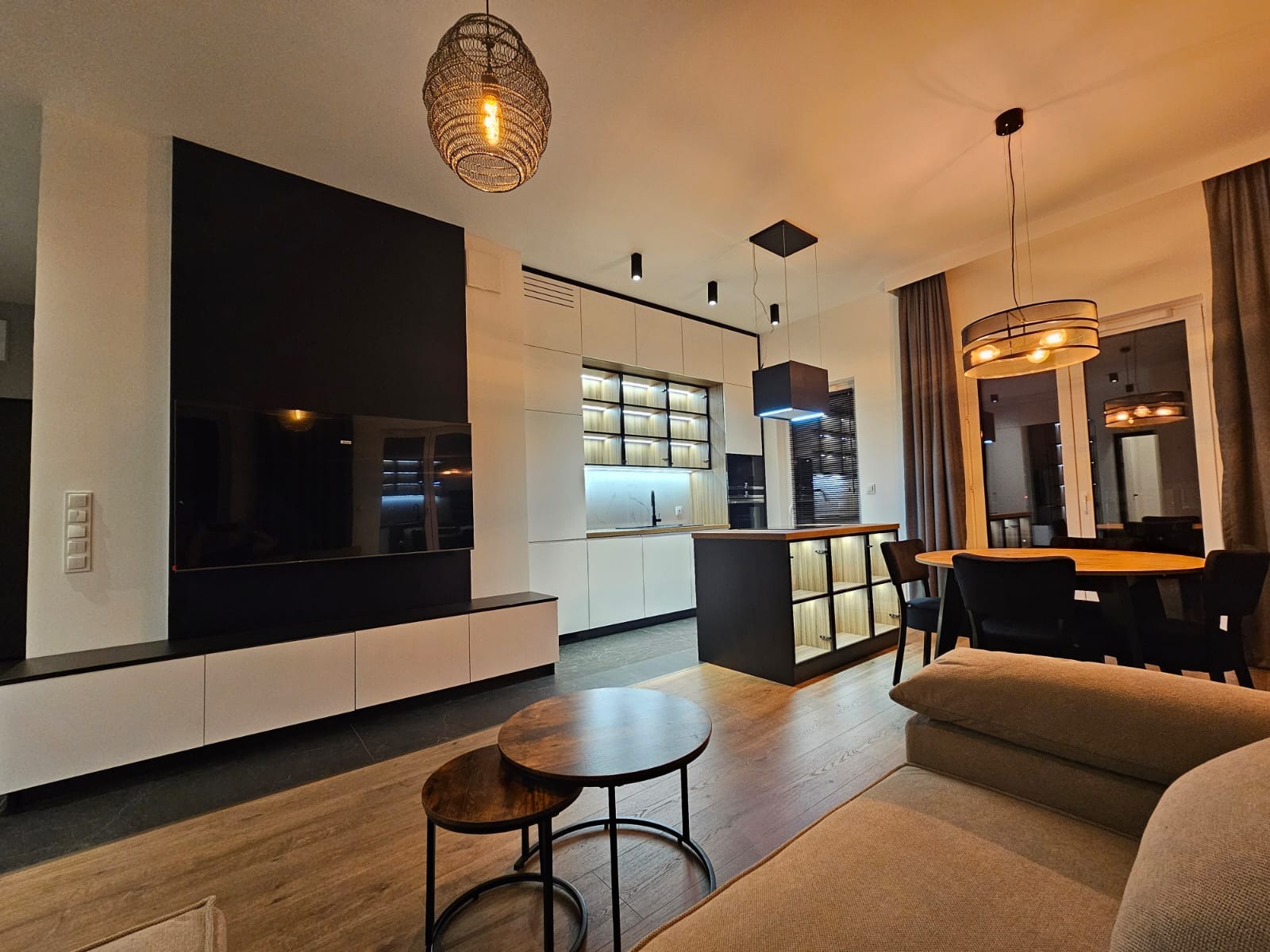 Nowoczesne wnętrze mieszkania z otwartą kuchnią, wyspą kuchenną, drewnianym stołem i elementami czerni, oświetlone designerskimi lampami wiszącymi.